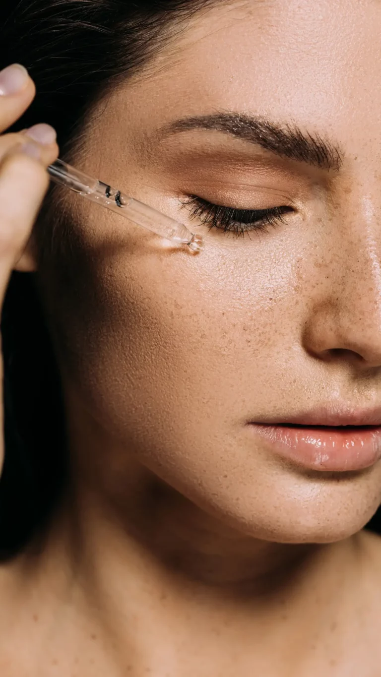 Close-up side view of a woman's face, focusing on her cheek, as a dropper dispenses a clear serum onto her skin. The woman's eyes are closed, showcasing long eyelashes, expertly applied eye makeup, and a clear complexion with freckles. The image suggests a moment of skincare treatment or beauty routine.