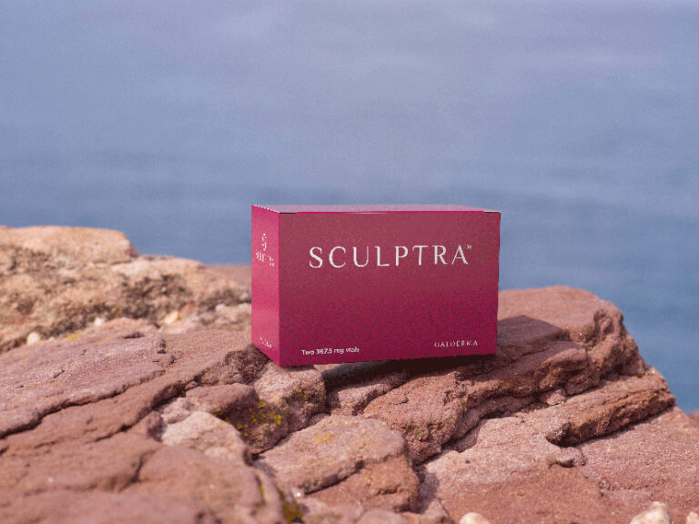 A maroon-colored box of Sculptra, a cosmetic injectable product, sits on rocky terrain overlooking a serene body of water in the background. The box features white text with the brand name 'Sculptra' prominently displayed, and it is positioned in a natural, outdoor setting, giving a sense of tranquility and beauty.