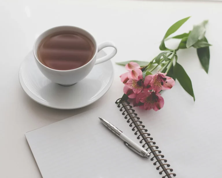 White cup of tea next to a spiral notebook, silver pen, and pink flowers on a white surface.