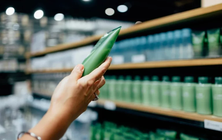 A person holds a green skincare product tube while shopping in a store. Shelves in the background display rows of similar green bottles, suggesting a variety of skincare products. The scene is slightly blurred, emphasizing the product in the person's hand.